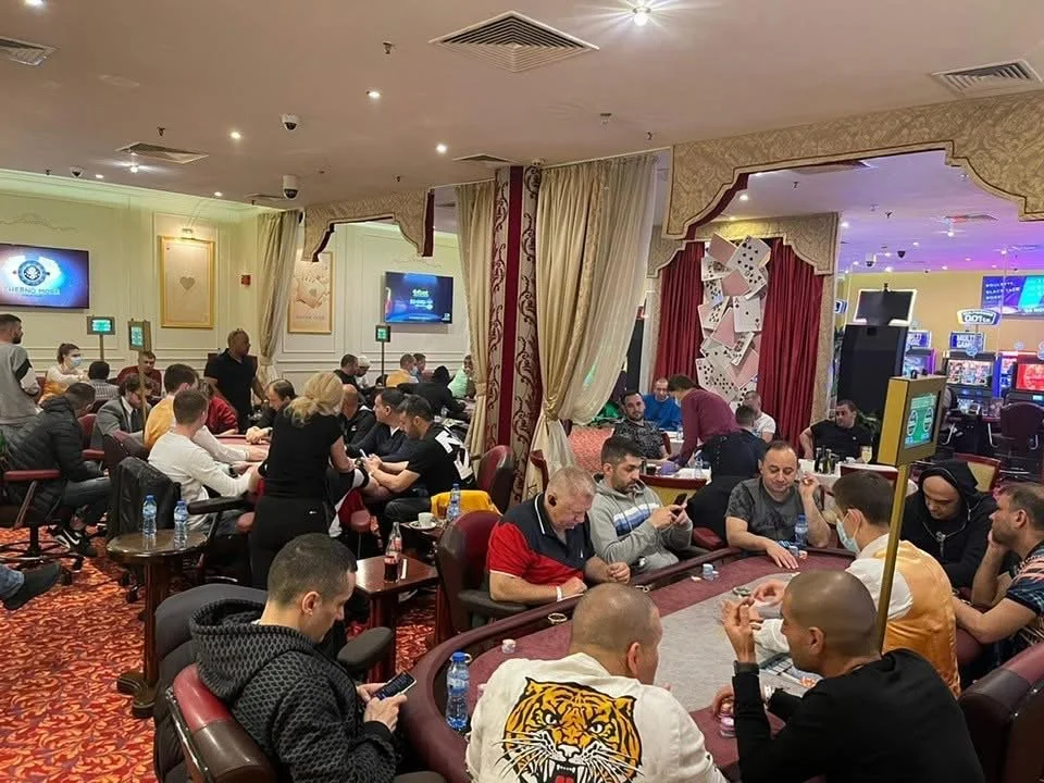 People playing poker at casino tables with slot machines visible in the background, in a decorated indoor setting.