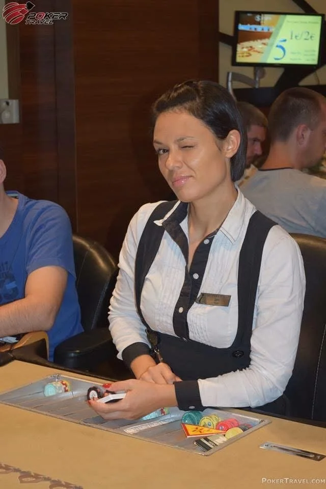 A waitress at a poker table winks and holds poker chips, wearing a black and white uniform, in a casino setting.