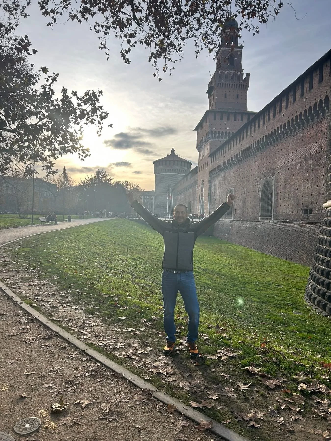 A man wearing a gray jacket and blue jeans with outstretched arms standing on a grassy area near a historic brick castle wall with a tower. The sky is cloudy with the sun setting behind trees.