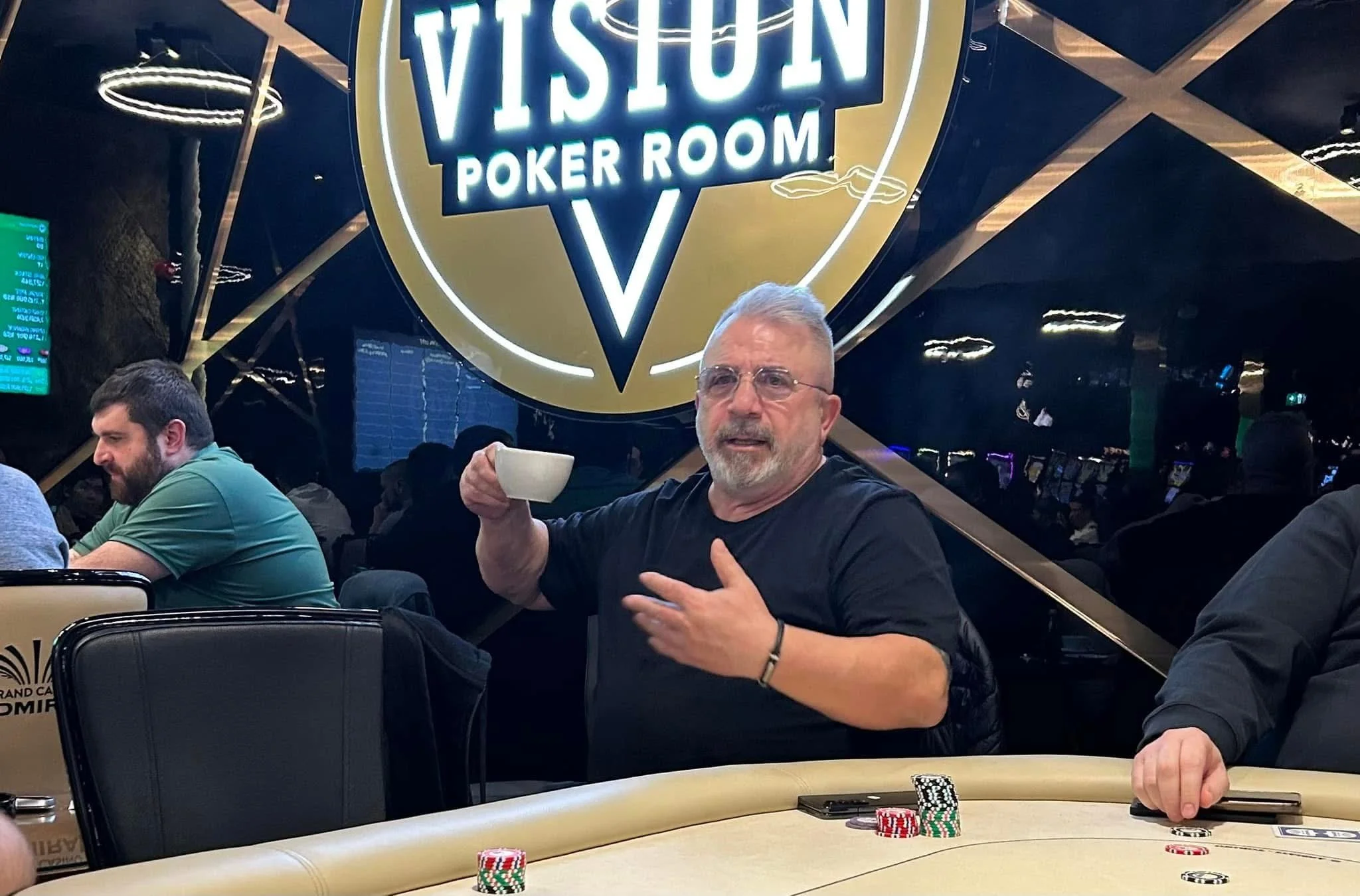 A man with gray hair and glasses holding a white cup at a poker table inside the Vision Poker Room. Several stacks of poker chips are on the table. In the background, a neon sign reads 'Vision Poker Room' and there are other people seated at the tabl