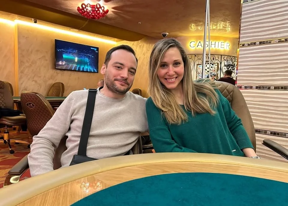 A man and a woman sitting at a poker table in a casino, smiling at the camera. The background includes a TV, a sign that says 'CASHIER,' and casino decor.