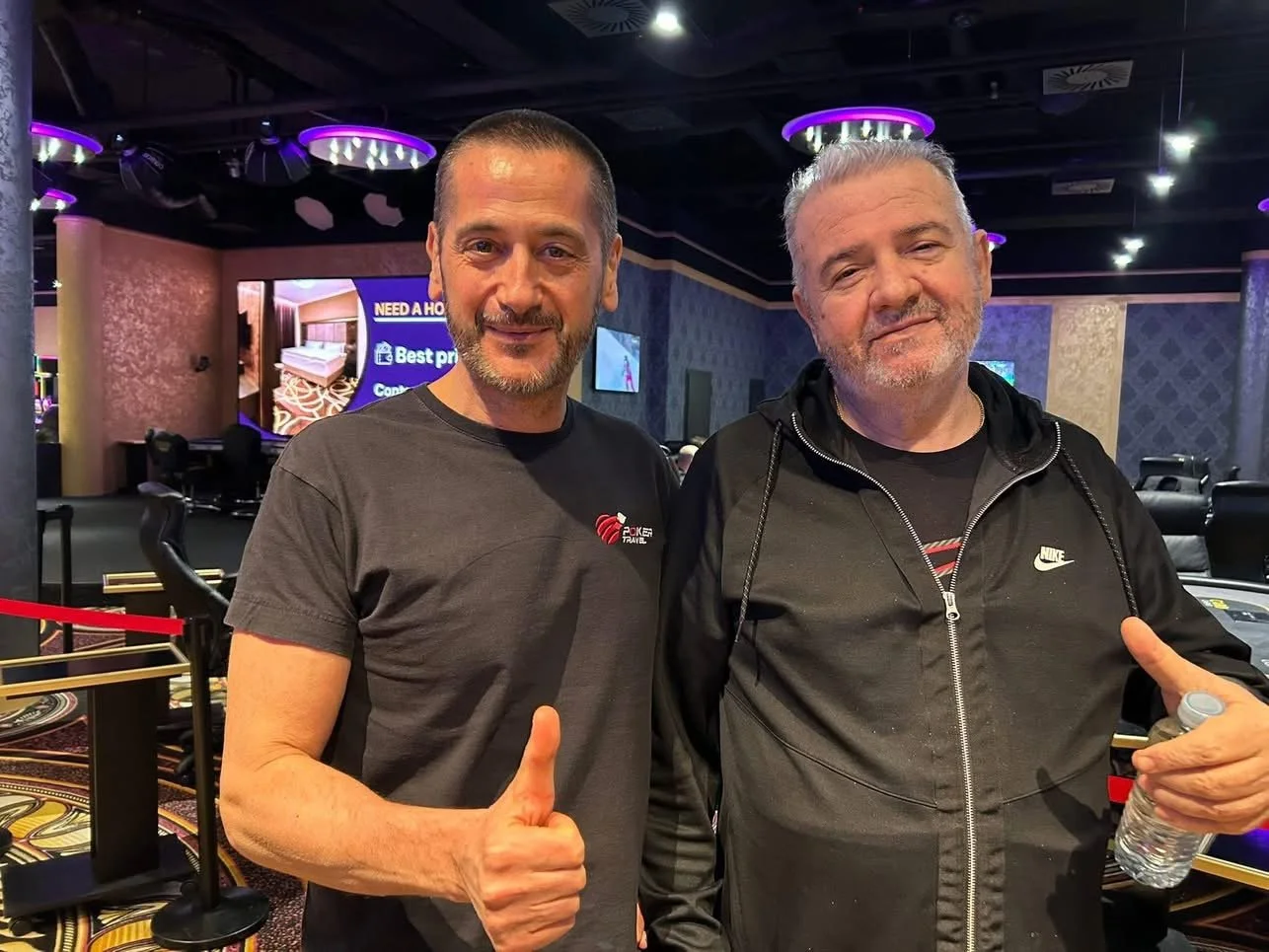 Two men in a casino or gaming room, one giving a thumbs-up and the other holding a water bottle, both smiling.
