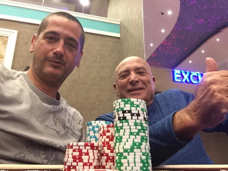 Two men sitting at a table with poker chips stacked in front of them, one giving a thumbs-up and both smiling, inside a casino or poker room.