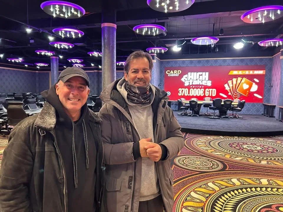 Two men standing inside a casino with poker tables in the background. They are smiling and wearing jackets, with purple lighting fixtures on the ceiling and a large screen displaying a poker tournament prize of 370,000 euros.
