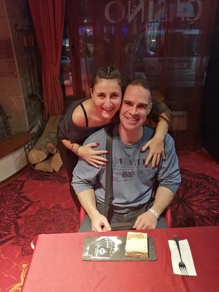 A woman and a man sitting at a restaurant table, smiling for the camera. The woman is hugging the man over his shoulders. There is a small piece of cake on the table in front of him.