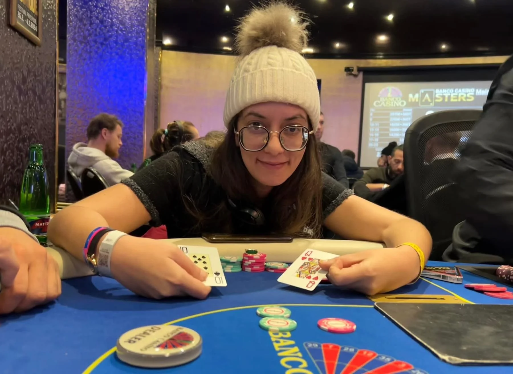 A woman with glasses and a knitted beanie smiling at a poker table during a casino event, with poker chips, playing cards, and other players visible in the background.