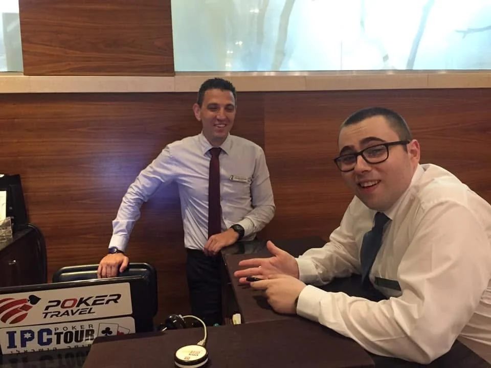 Two men in dress shirts and ties at a poker tournament table, smiling and interacting. One is standing behind the table with a hand on a chair, the other is sitting, gesturing toward the camera.