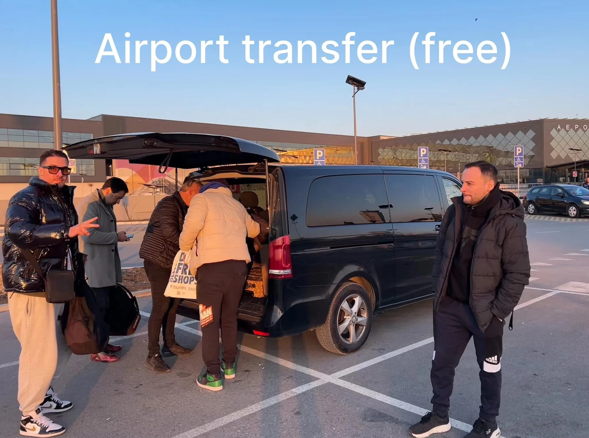 People standing around a black vehicle at an outdoor airport parking lot, with the airport terminal building in the background and a clear sky overhead.