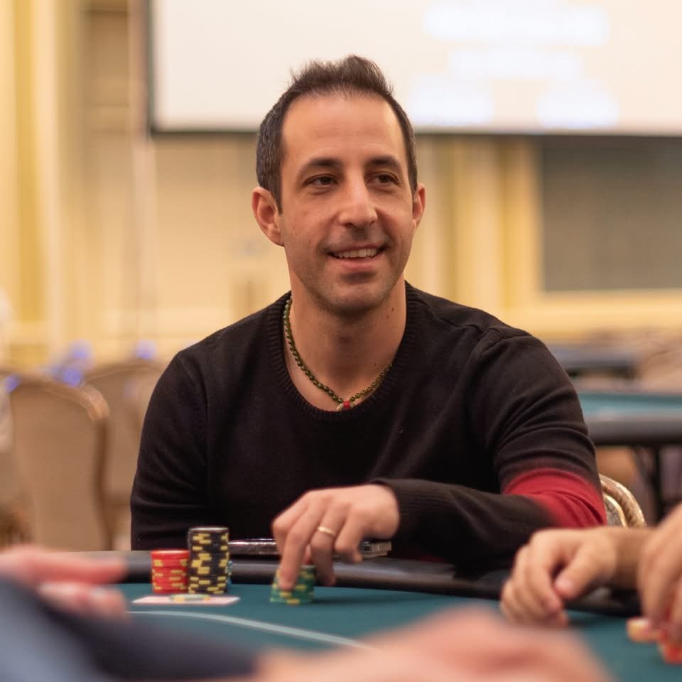A man smiling while participating in a poker game at a casino or poker room, with poker chips and cards on the table.
