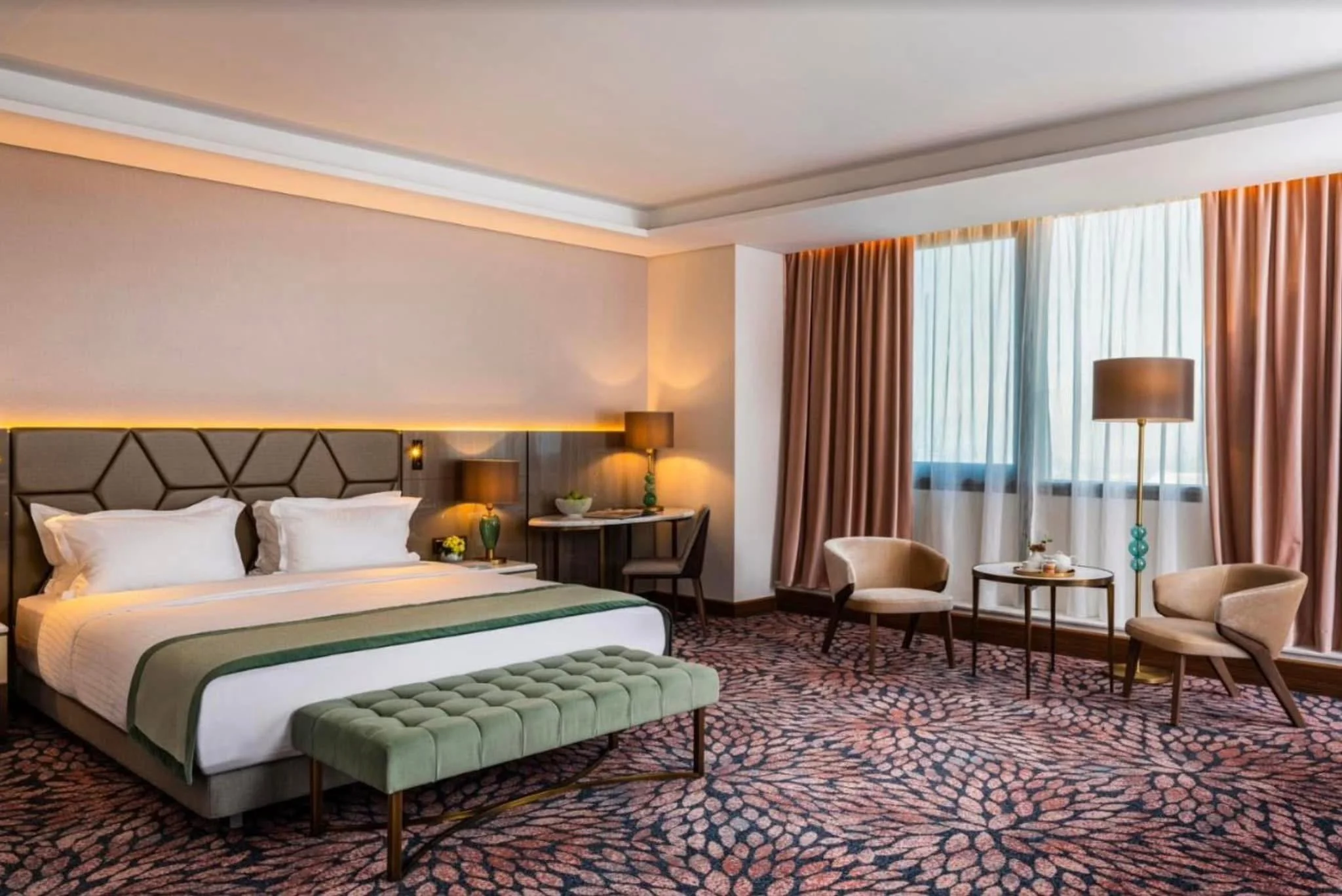 A hotel room with a king-sized bed, nightstands, lamps, a small seating area with chairs and a table, and large windows with curtains.