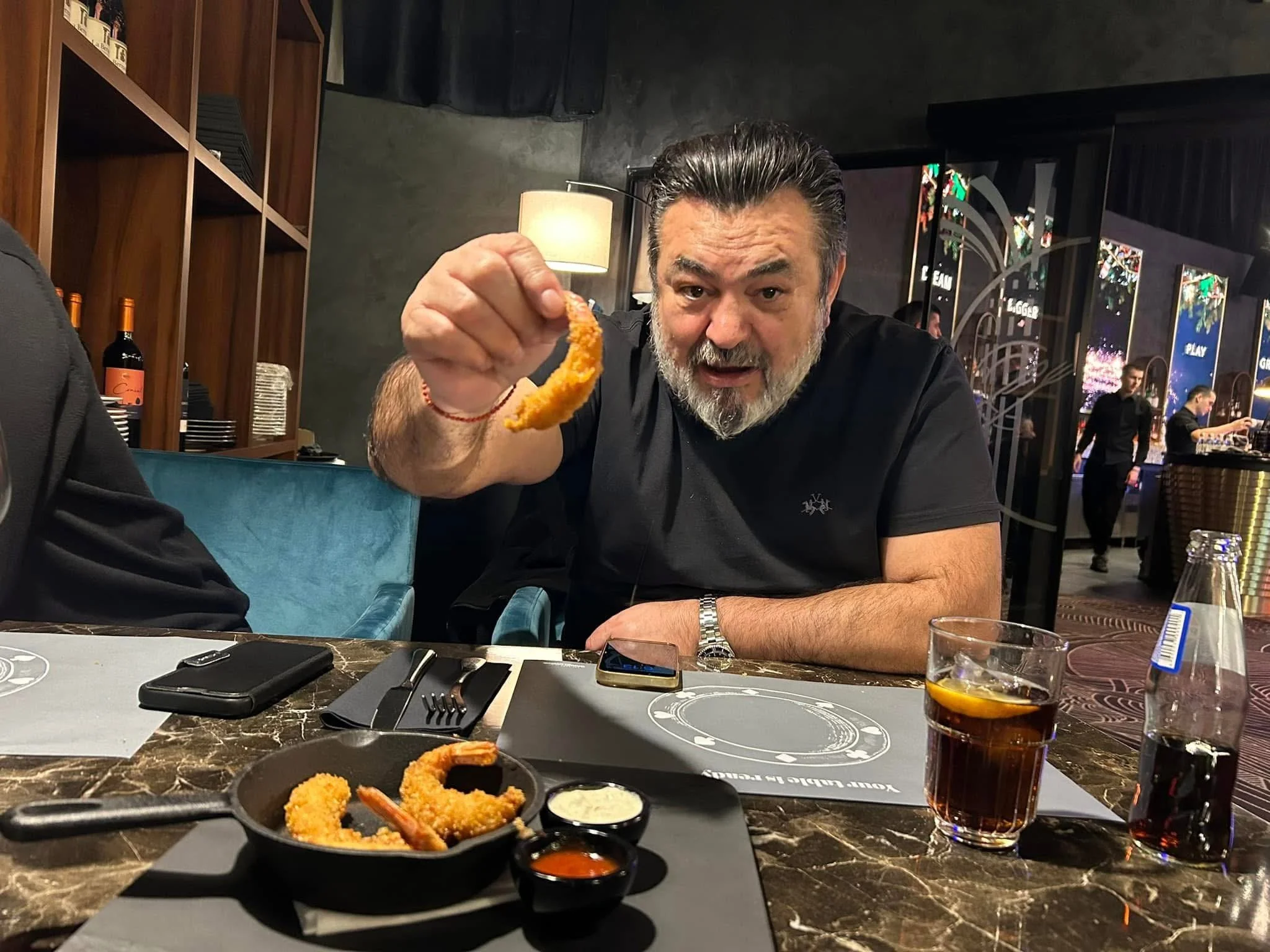 A man with a gray beard and dark hair holding a fried shrimp in a restaurant. There are plates of fried shrimp with dipping sauce and glasses of soda on the table, with a busy restaurant background.