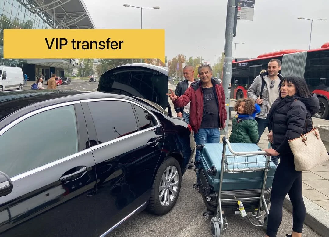 Travelers waiting for VIP transfer at airport with luggage cart, near parked black car and red buses.