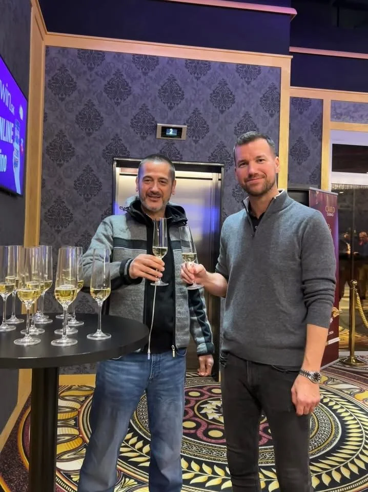 Two men standing inside a hotel or casino, holding glasses of champagne, with a table of additional champagne glasses nearby, in a decorated, upscale setting.