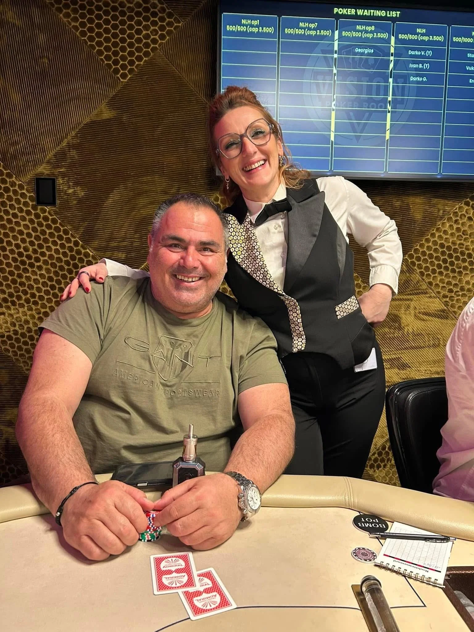 Two people smiling at a poker game, with poker chips, playing cards, and a poker table in front of them. One man is sitting and holding chips, while the woman stands behind him, dressed as a dealer.