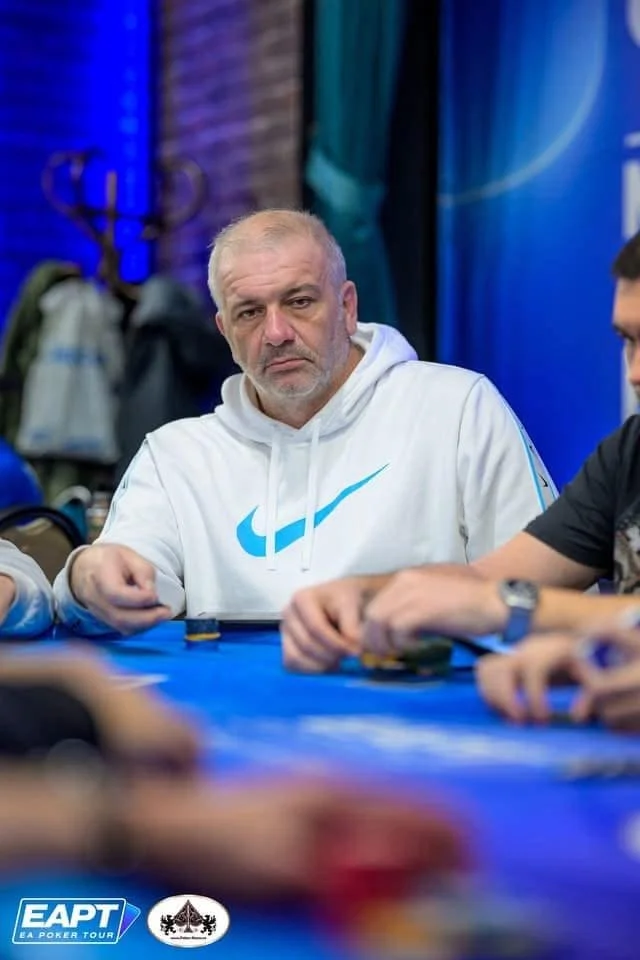 A middle-aged man with gray hair and beard sitting at a poker table with a serious expression, wearing a white hoodie with a blue Nike logo, surrounded by other players, in an indoor setting with poker chips and a blue table.
