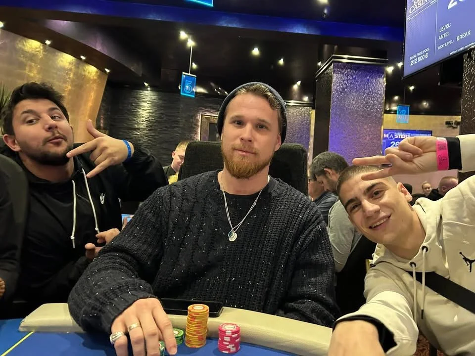 Three young men at a poker table in a casino, with poker chips and cards in front of them, smiling and making peace signs.