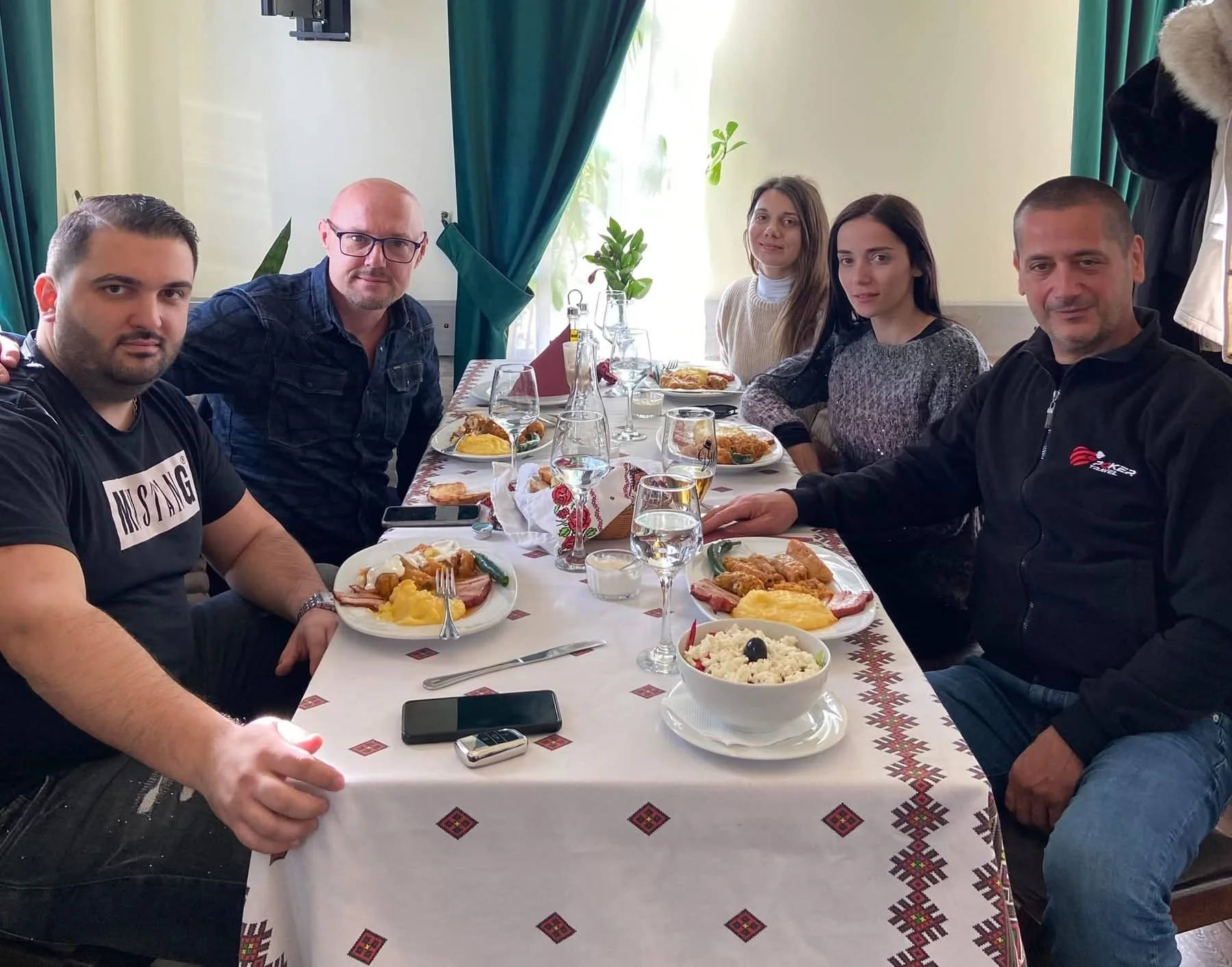 A group of six people sitting at a dining table with plates of food, glasses, and a bowl of rice with a black olive on top. The table is covered with a white tablecloth with a traditional pattern. The background shows a window with green curtains and