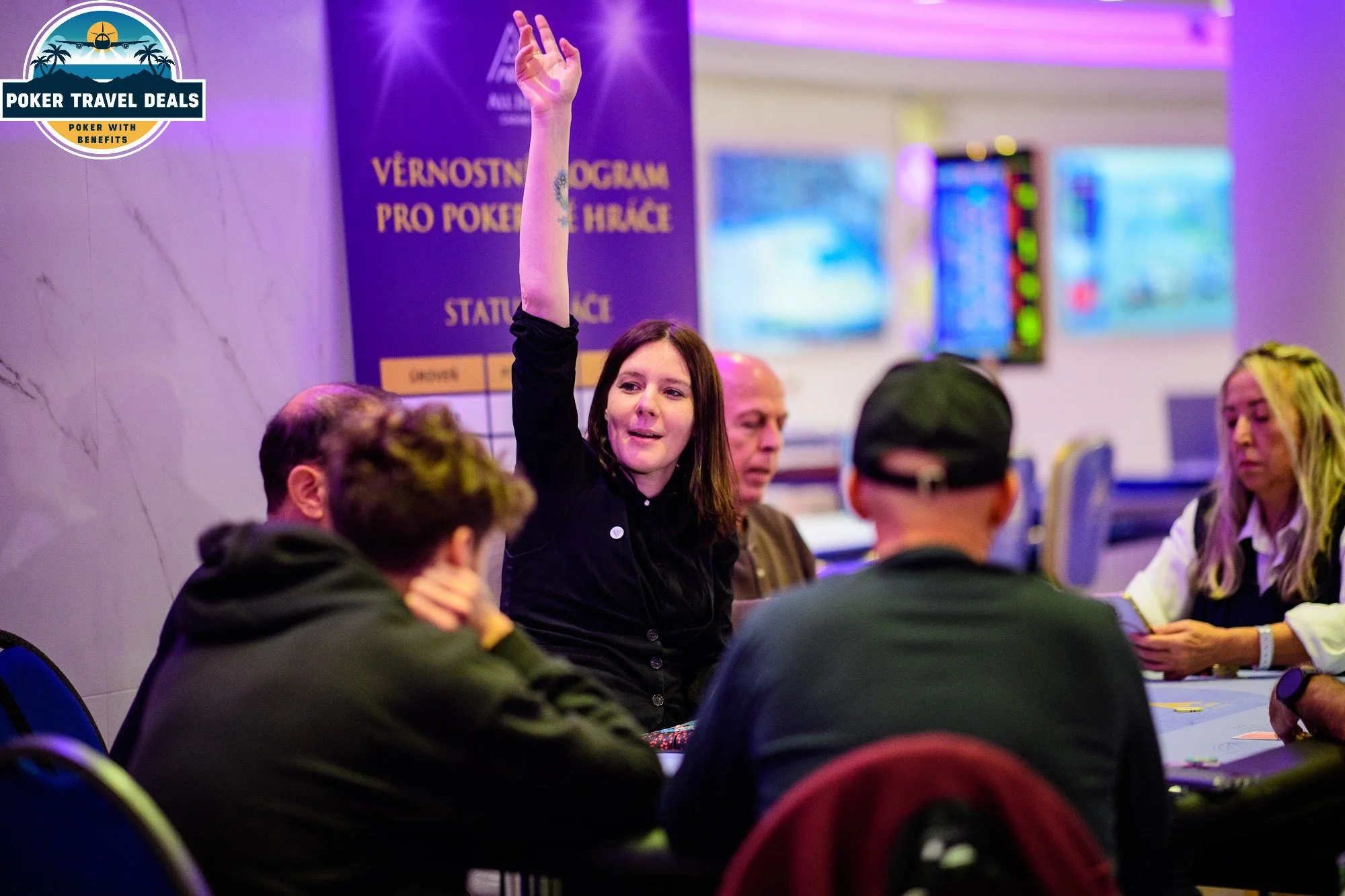 A woman with dark hair raising her hand at a poker event, surrounded by people sitting around a poker table in a room with purple lighting.