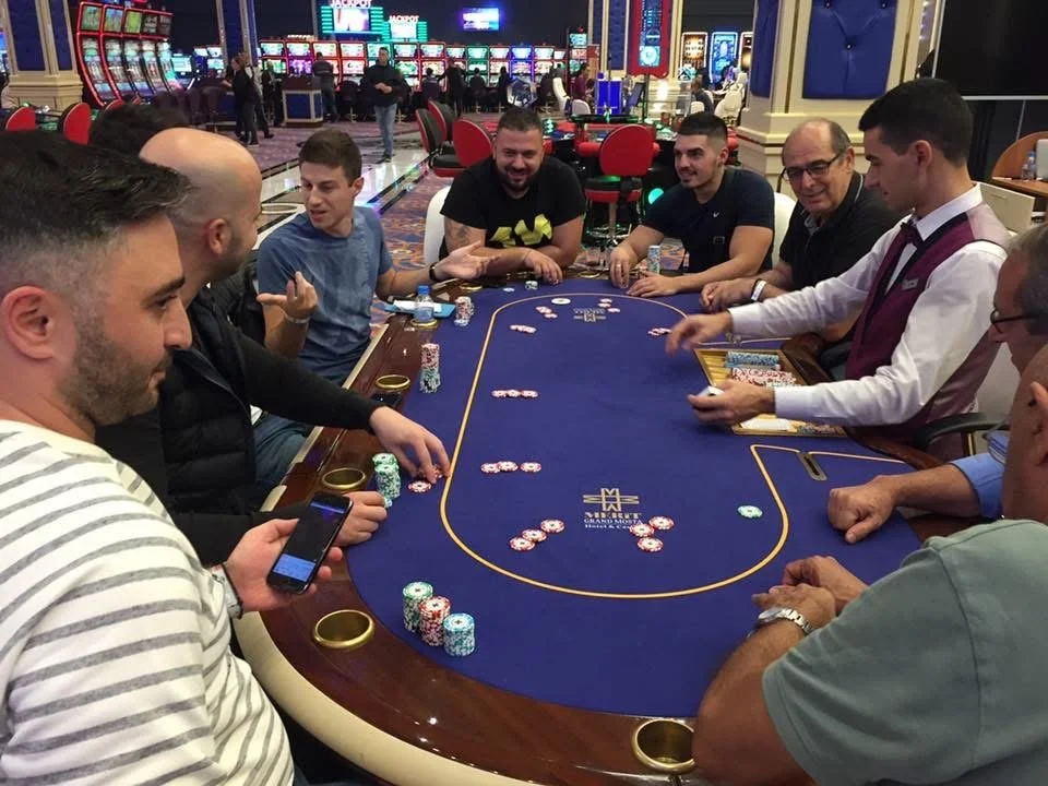 A group of people playing poker at a casino table with chips and cards, with slot machines and bright lights in the background.