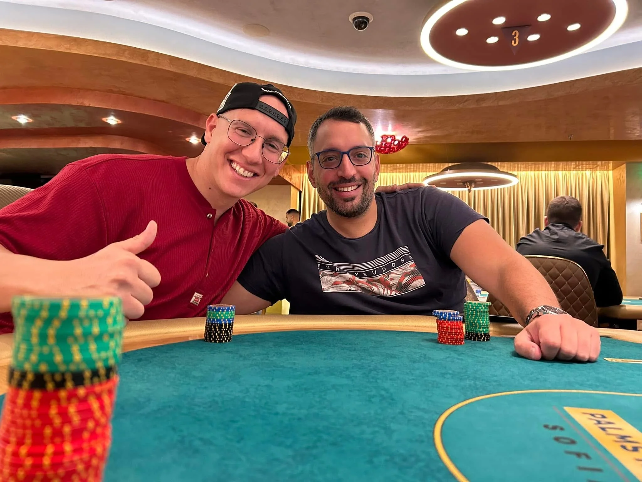 Two men smiling sitting at a poker table, with poker chips in the foreground, in a casino or poker room.