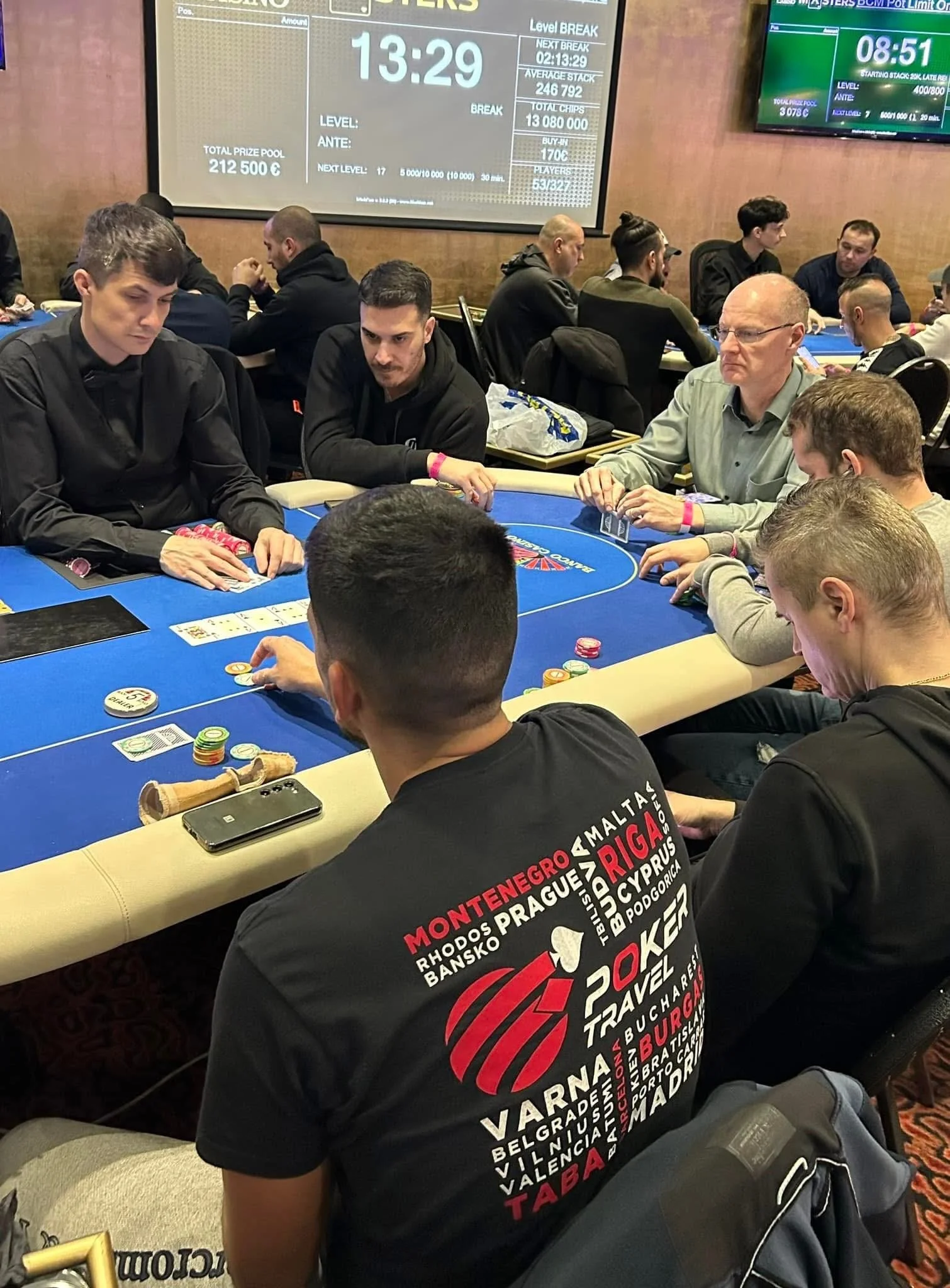 A group of people sitting at a poker table playing card game, with chips and other game accessories on the table. Large screens in the background display poker game stats and timer.