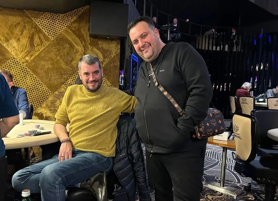 Two men smiling in a casino, one seated with a yellow sweater and the other standing with a black jacket, surrounded by poker chips and gaming tables.