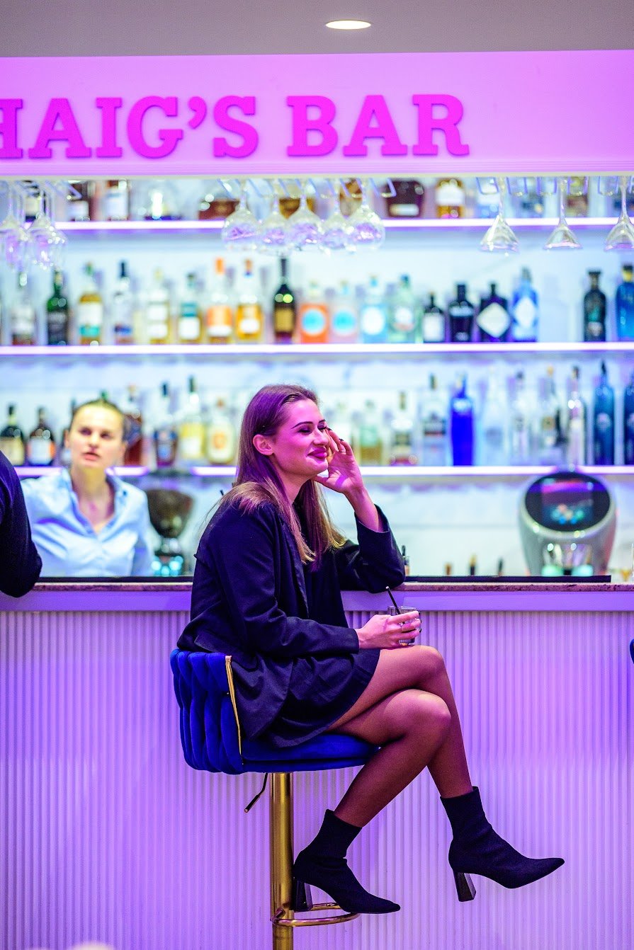 A woman with long hair sitting on a blue barstool at a bar called 'HAIG'S BAR' with a bar counter and shelves of liquor bottles in the background. She is wearing a black blazer, black skirt, and black high-heeled boots, holding a drink, and smiling. 