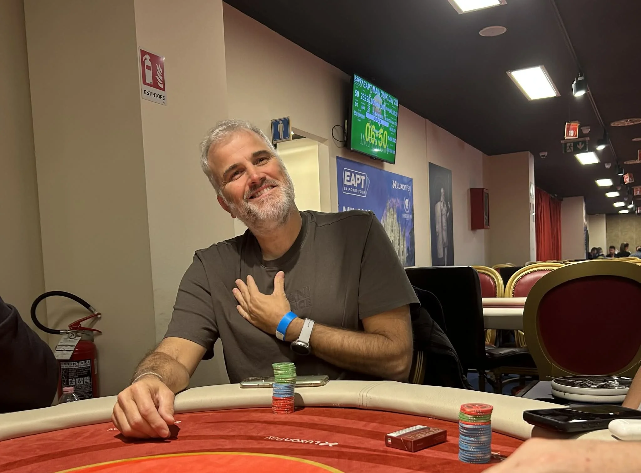 A man with gray hair and a beard smiling and sitting at a poker table with poker chips in front of him. He has a wristband on his right wrist and is wearing a dark shirt. The background shows a television screen displaying information and several peo