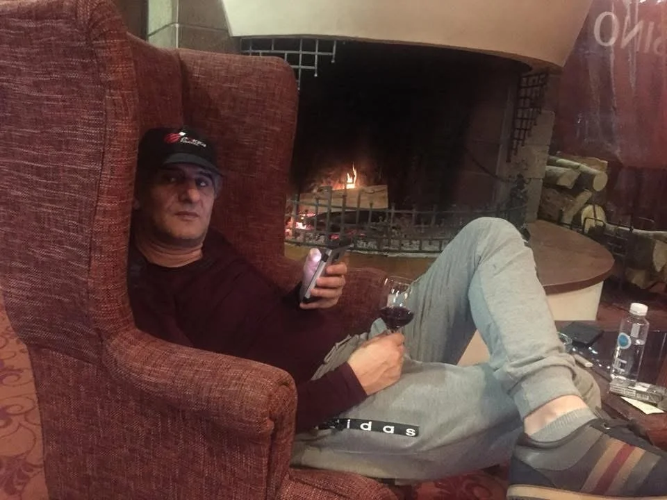 Man sitting in a large chair by a fireplace, holding a remote control in one hand and a glass of red wine in the other.