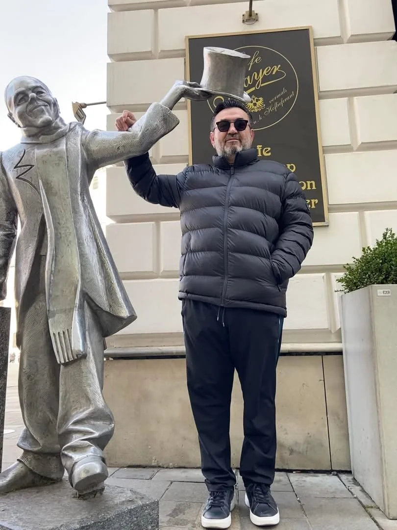 A man in a black puffer jacket and sunglasses standing next to a silver statue of a man holding a hat. The statue's hand is outstretched, holding the hat over the man's head. They are outside a building with a sign in the background reading "Cafe" an