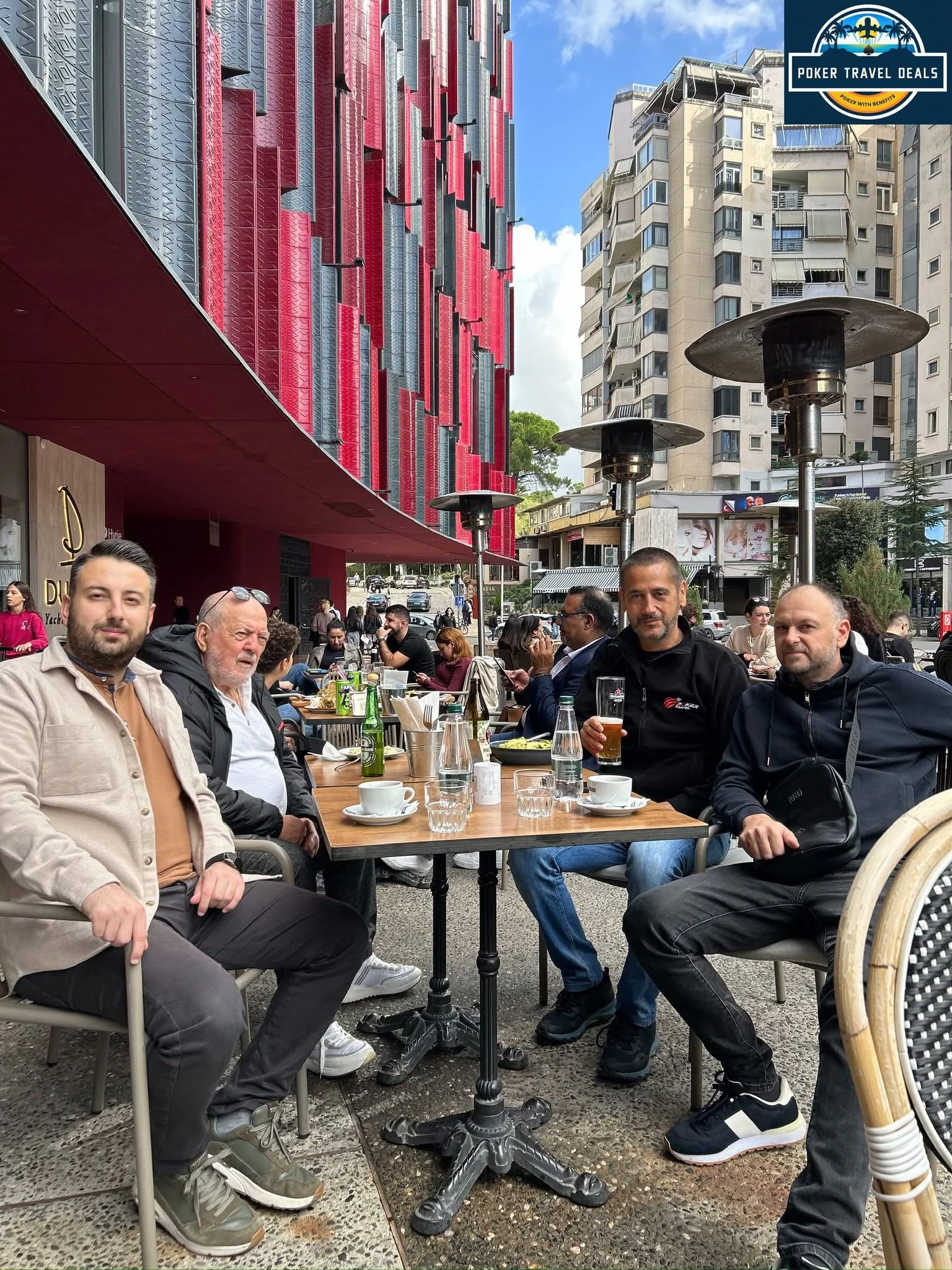 A group of people sitting at a large outdoor café table with food and drinks, in an urban setting with tall buildings and heaters in the background, under a partly cloudy sky.