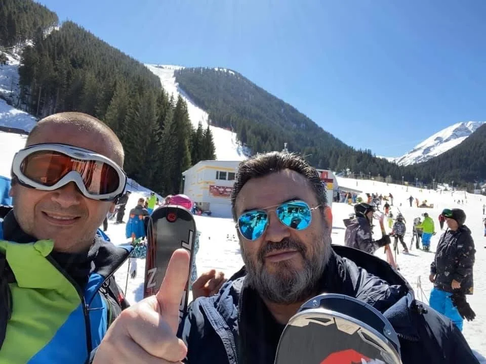 Two men in ski gear taking a selfie on a snowy mountain, with other skiers and snow-covered slopes in the background.
