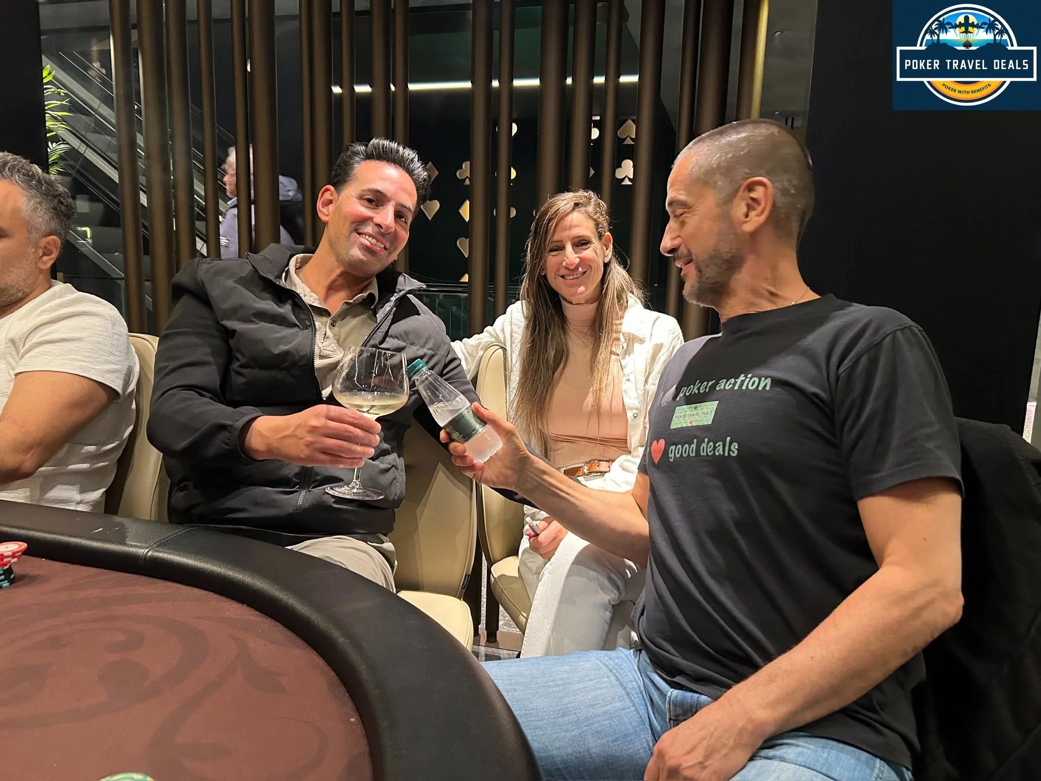 Four people sitting at a poker table, two men and two women, smiling and chatting. One man is holding a bottle of water and a wine glass, and is passing a fanned card to another man. The background features decorative vertical gold rods, and a sign i