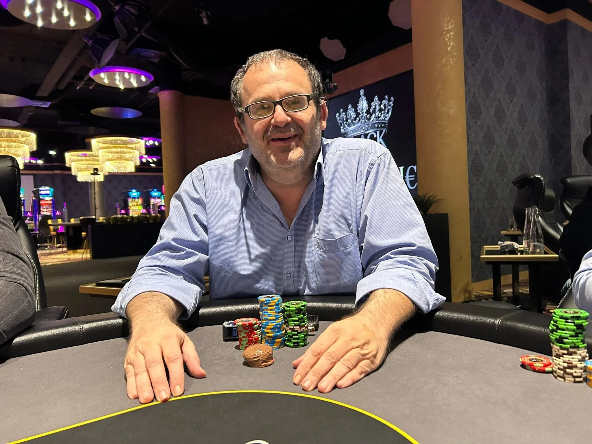 A man with glasses and a beard smiling at a poker table, surrounded by colorful poker chips, in a casino or gaming room with purple and gold decor and light fixtures.