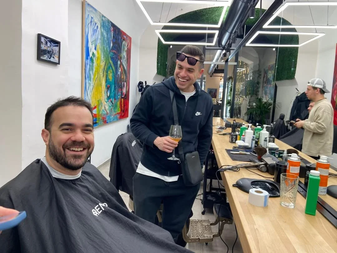 Two men smiling in a barbershop, one seated in a barber chair and the other standing with sunglasses on his head, holding a drink, with hairdressing tools and products on the counter.