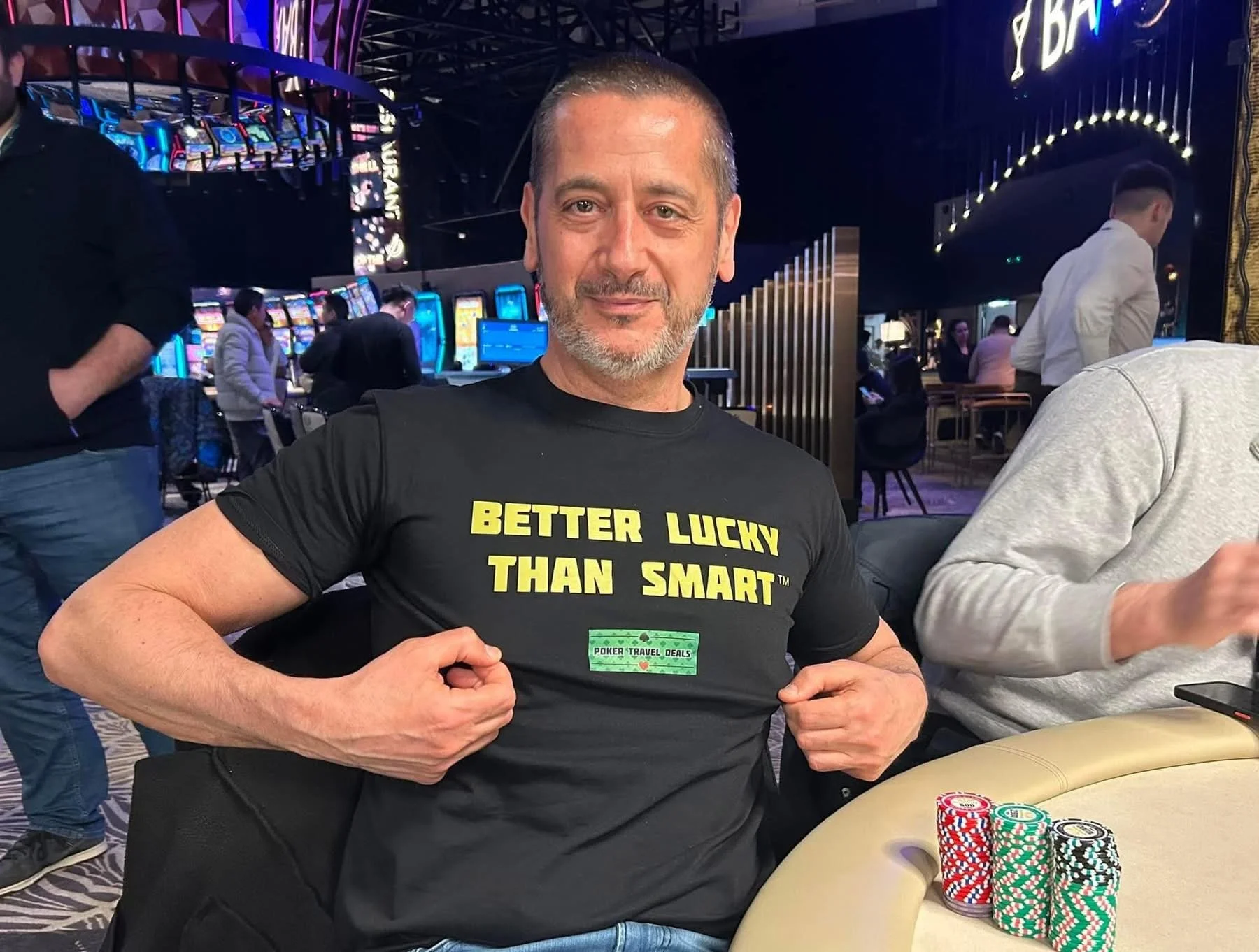 A man sitting at a poker table in a casino, flexing his arms, wearing a black T-shirt that says 'Better Lucky Than Smart' with poker chips in front of him on the table. The background shows slot machines and other people playing.