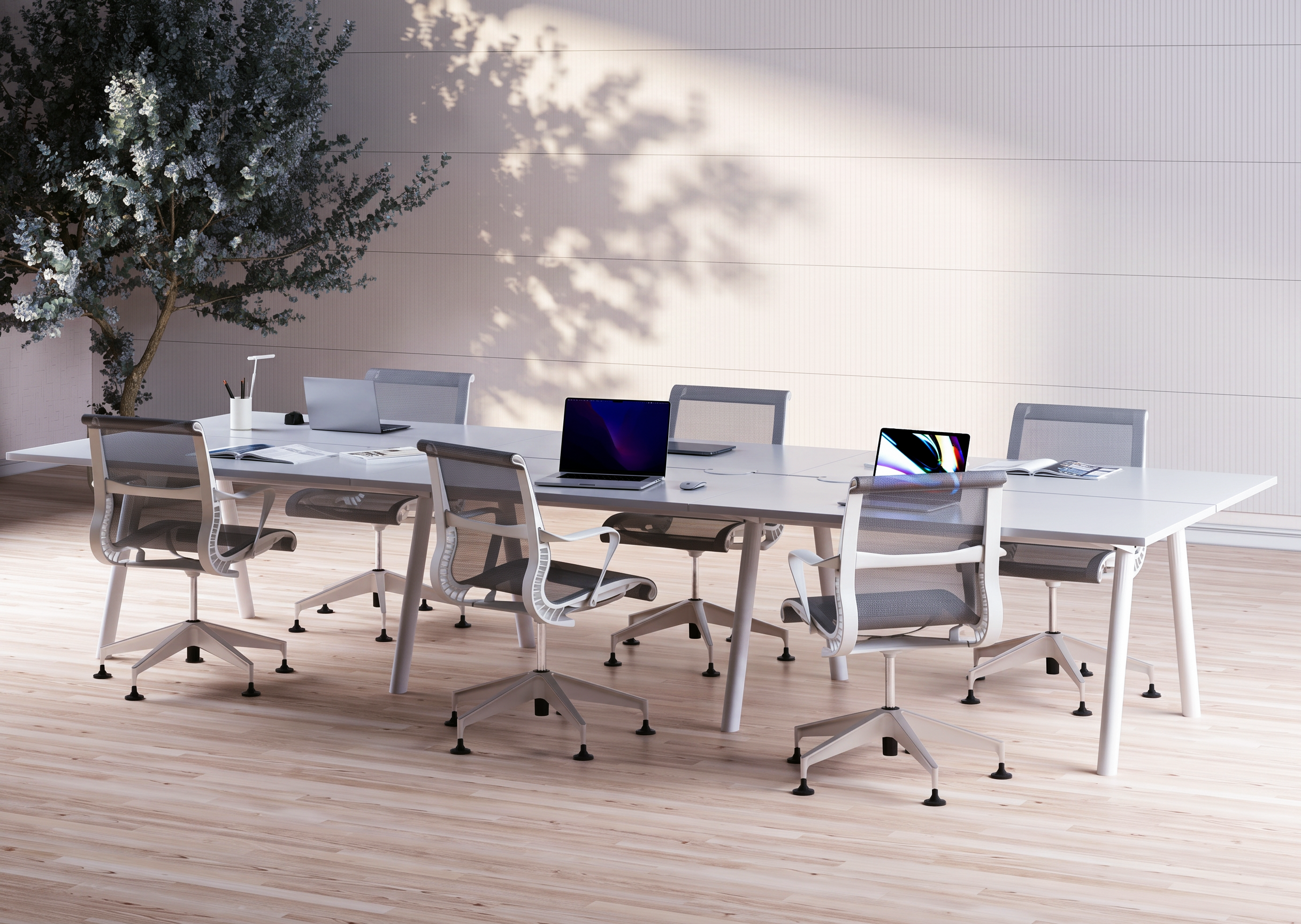 Modern conference room with a long white table, multiple office chairs, laptops, notebooks, and pens. A large tree with green leaves and white flowers is outside, casting shadows on the light gray wall inside.