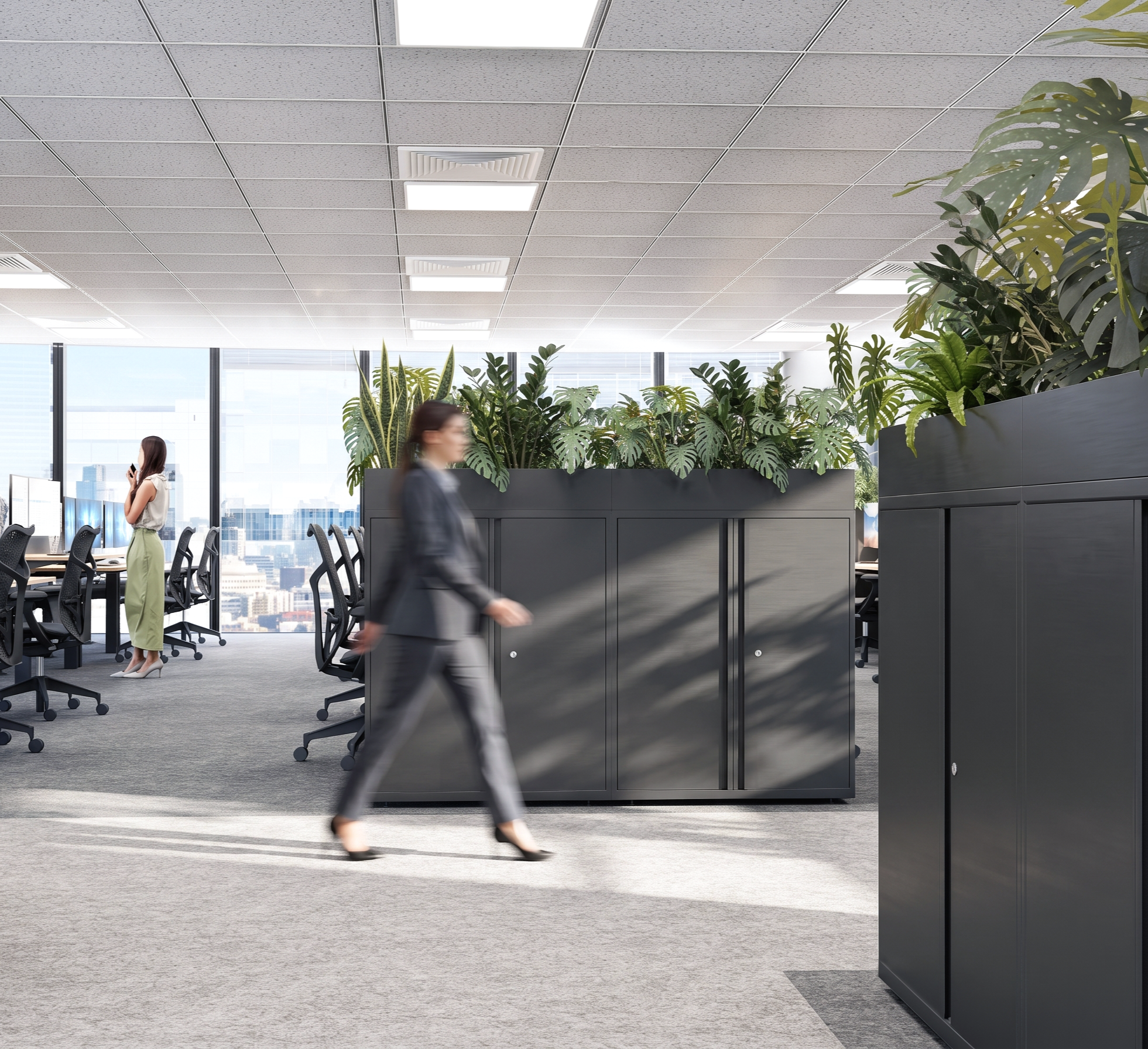 Office space with ceiling tiles, fluorescent lights, large windows, and indoor plants. Two office workers are walking, one woman standing by her desk and another woman blurred in motion walking past a black storage cabinet.