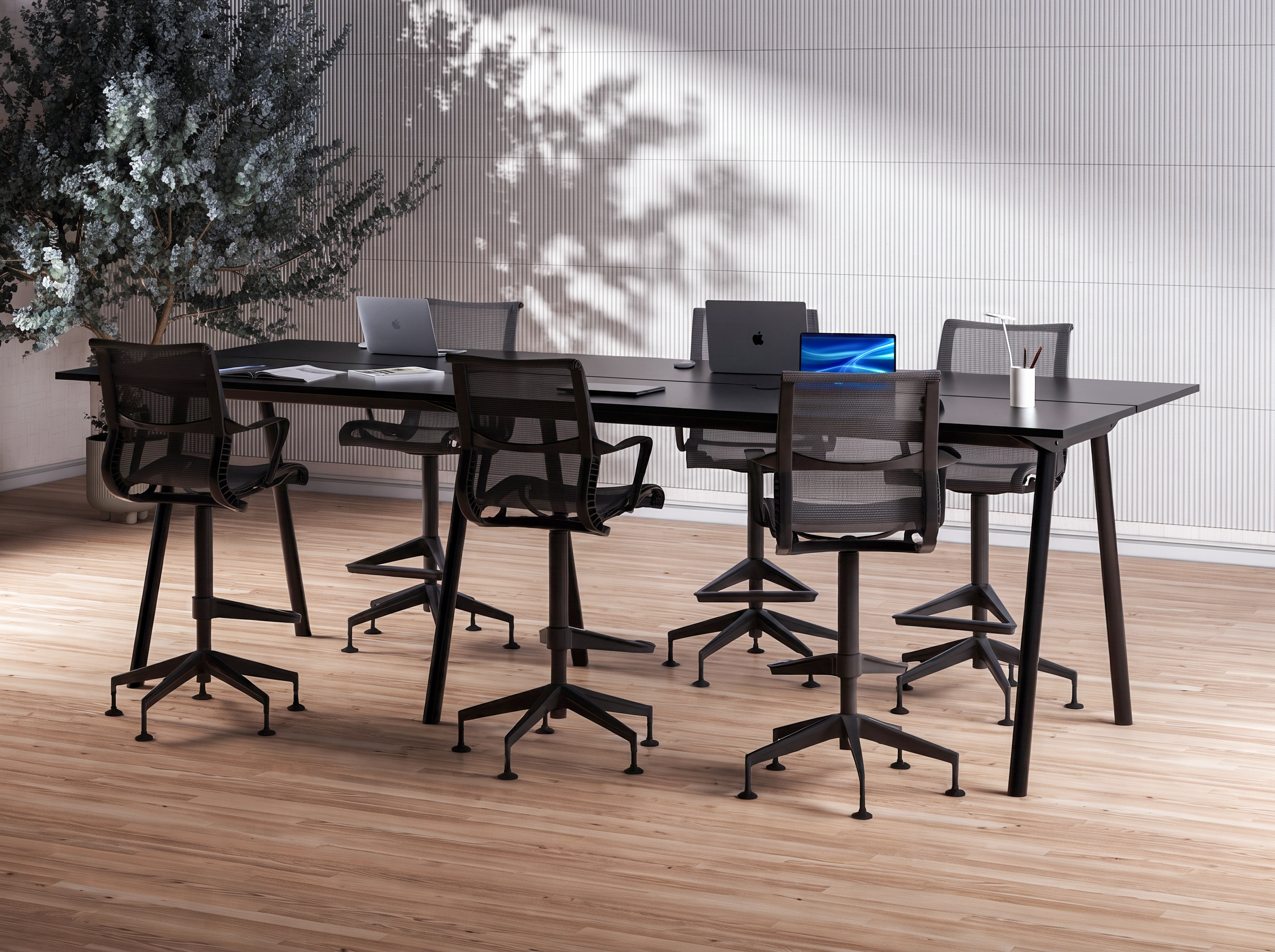 A modern office conference room with a long black table, six black mesh chairs, three MacBook laptops, an open notebook, and a desk lamp. There is a large indoor tree with textured leaves on the left side and a striped white wall on the right. The room has light wood flooring.