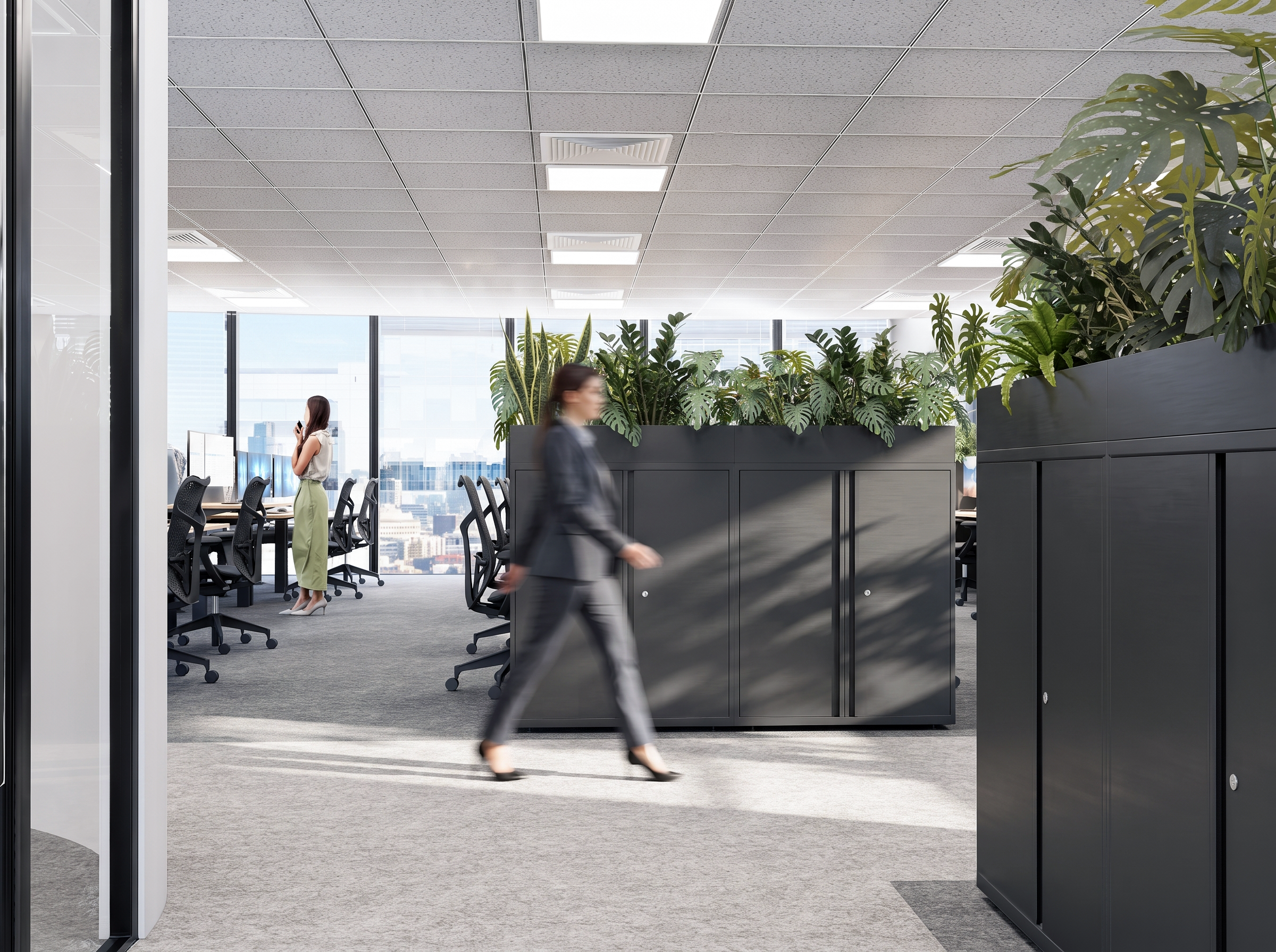 A modern office interior with high-rise city views, featuring workstations, black chairs, large green plants, and ceiling lights, with two women walking and a woman standing working on a computer.