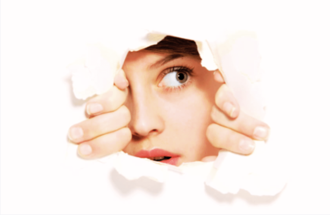Person peering through a torn hole in white paper with their face and eye visible. Curio identity image.