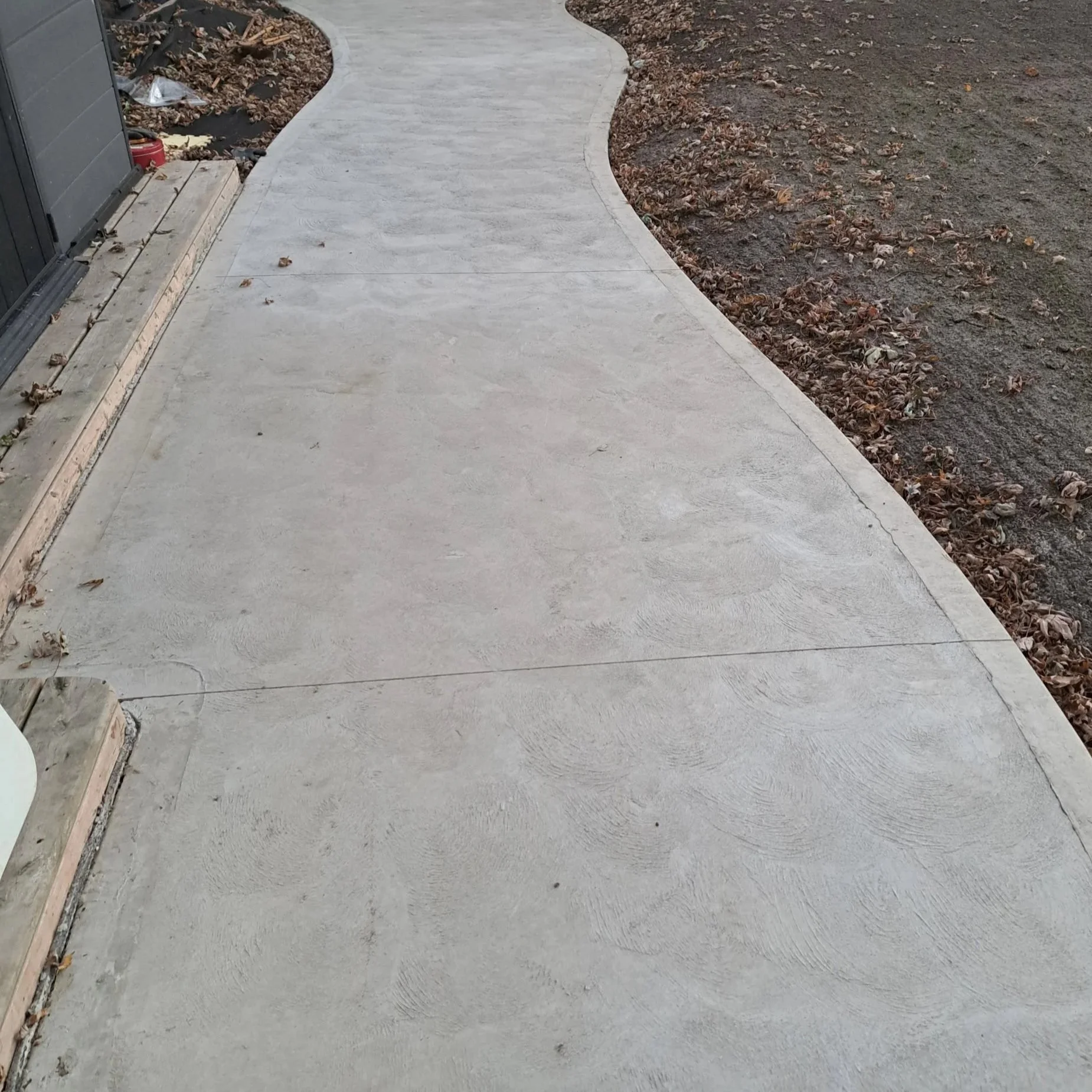 Powdered concrete sidewalk extending from the bottom left to the top right, with fallen leaves and a wooden border along the edge, next to a garden bed with dirt and debris on the left side and bare soil with scattered leaves on the right side.