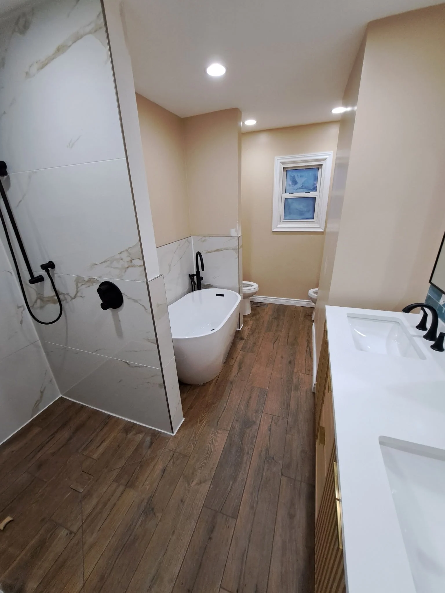 Modern bathroom with wood-look flooring, a white bathtub, a beige wall with a window, a vanity with a white sink, and black fixtures, with tiled walls