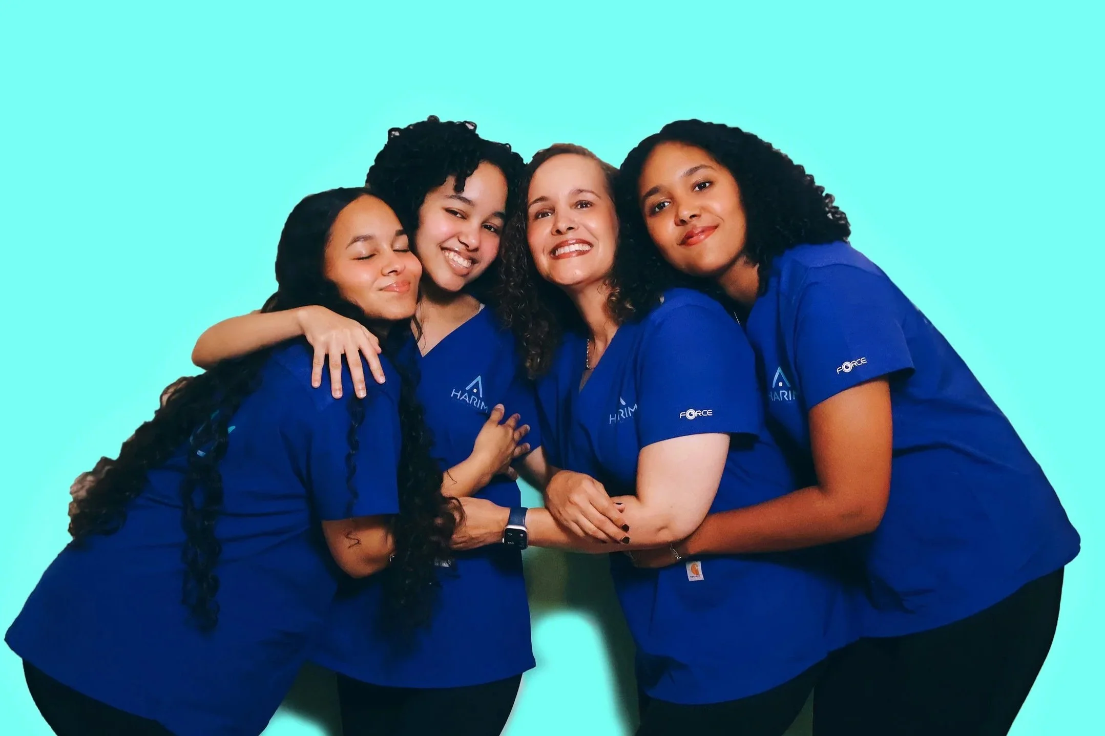 Four women hugging and smiling, wearing matching blue shirts against a light blue background.
