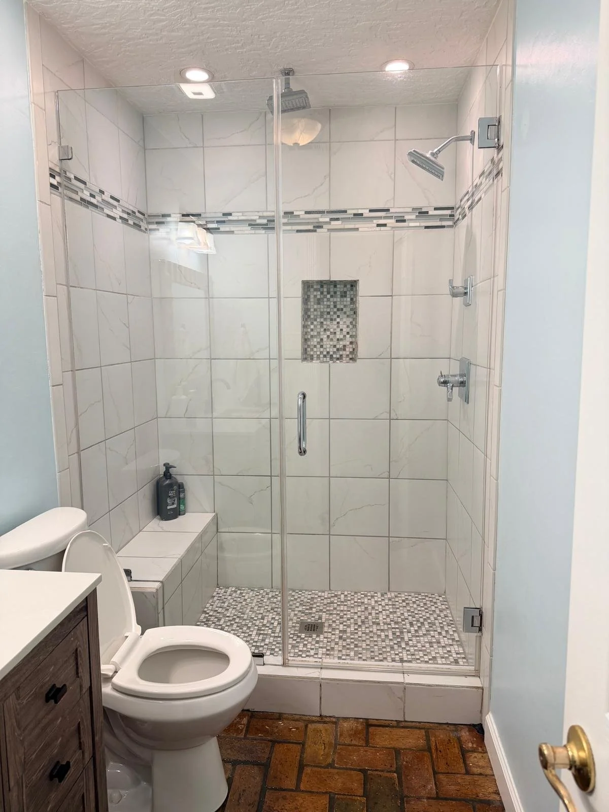 Bathroom with a tiled walk-in shower with a glass door, a built-in shelf, and multiple shower heads, next to a toilet and a small bathroom vanity.