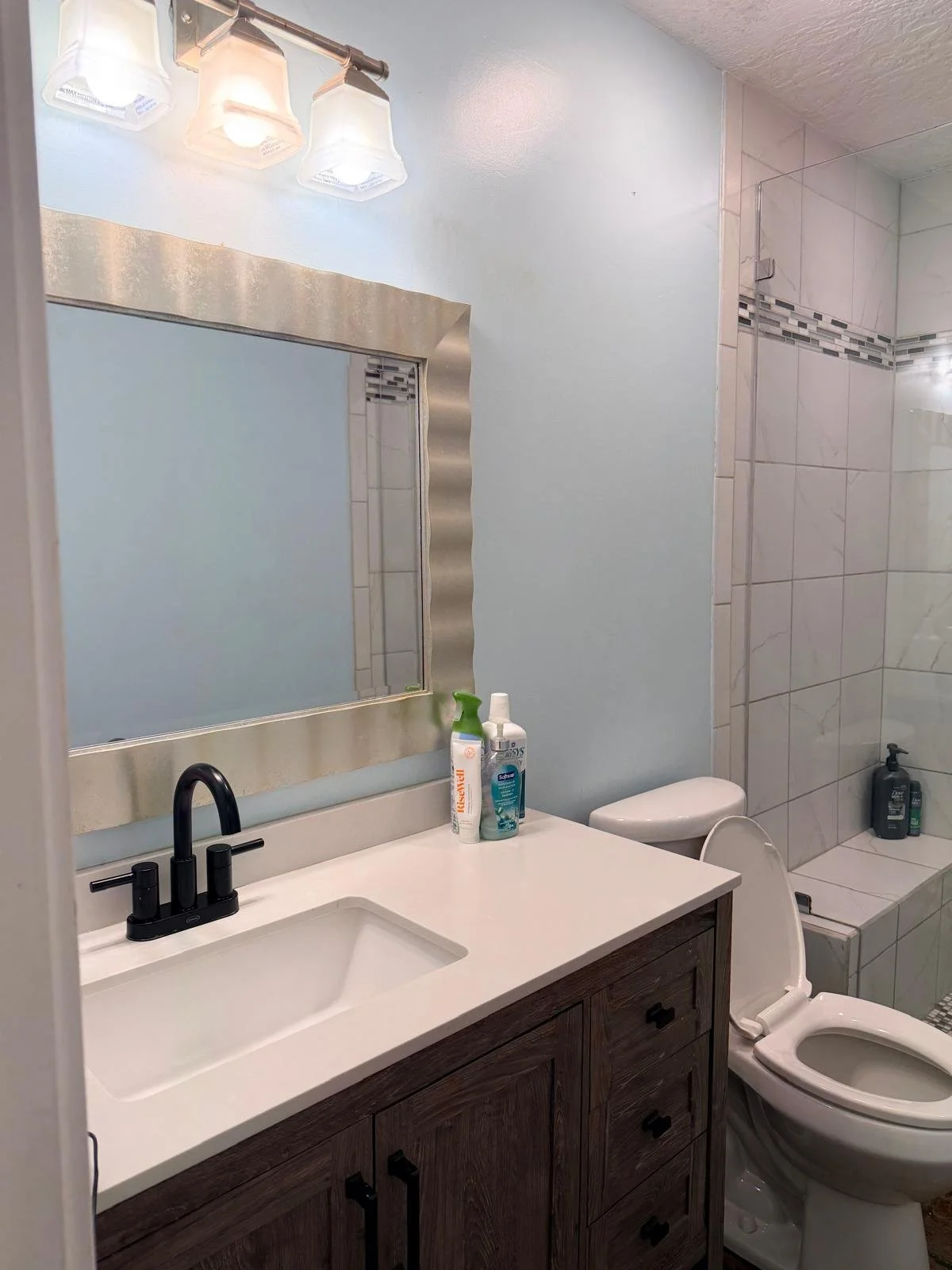 Bathroom with a mirror, sink with black faucet, toiletries, toilet, shower area with tiled wall, and a bottle of shampoo or soap on the shower ledge.