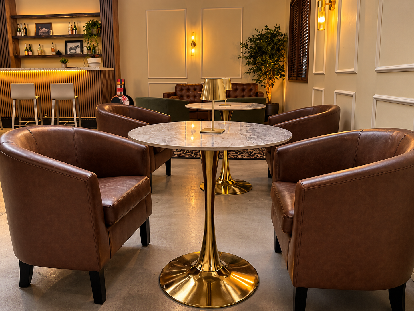 Cozy lounge area with a round marble table, four brown leather armchairs, and gold accents. In the background, there is a bar with bar stools, shelves with bottles, and decor, illuminated by warm lighting.