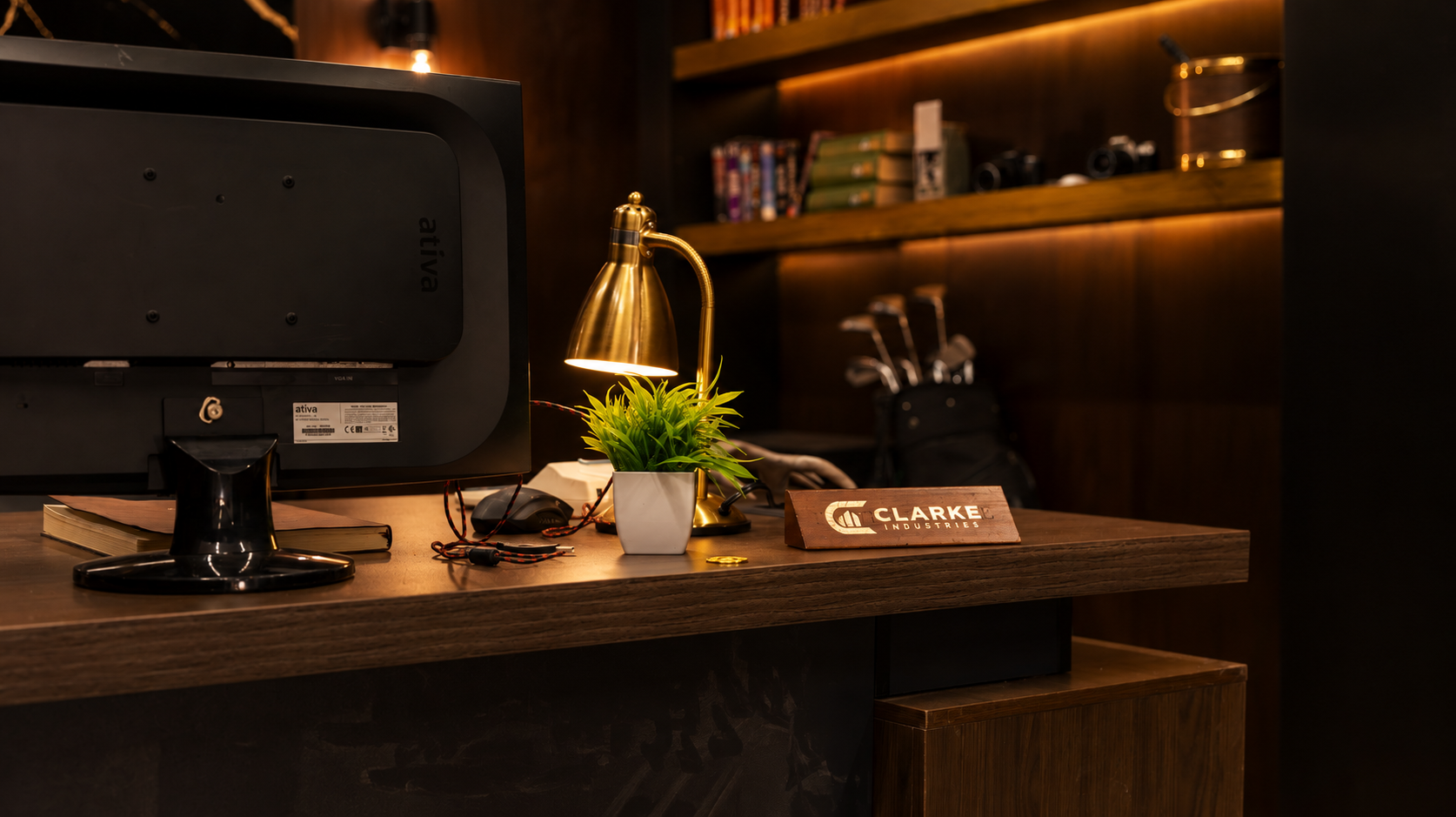 Office desk with a computer monitor, a brass desk lamp illuminating a potted plant, a note card with 'Clarke Industries' printed on it, and a pair of eyeglasses. In the background are wooden shelves with books and camera equipment.