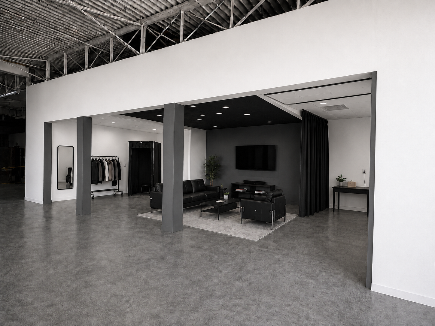 Modern lounge area with black leather sofas, wall-mounted TV, gray accent wall, and black curtains, adjacent to a clothing rack with jackets and coats, inside a spacious high-ceiling industrial-style space.