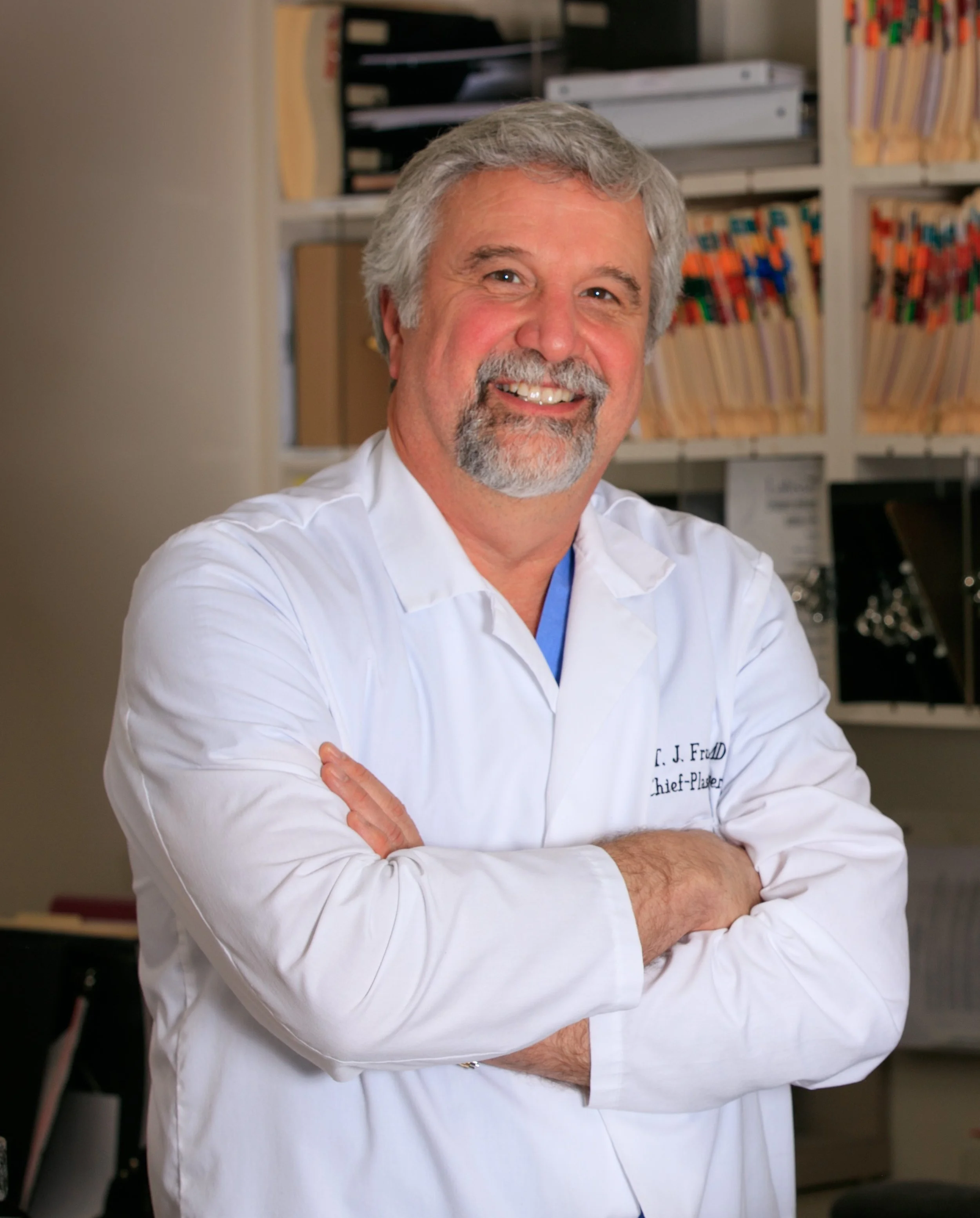 A smiling man with gray hair and facial hair wearing a white medical coat with blue shirt underneath, standing in front of bookshelves with organized files and folders in an office or clinic setting.